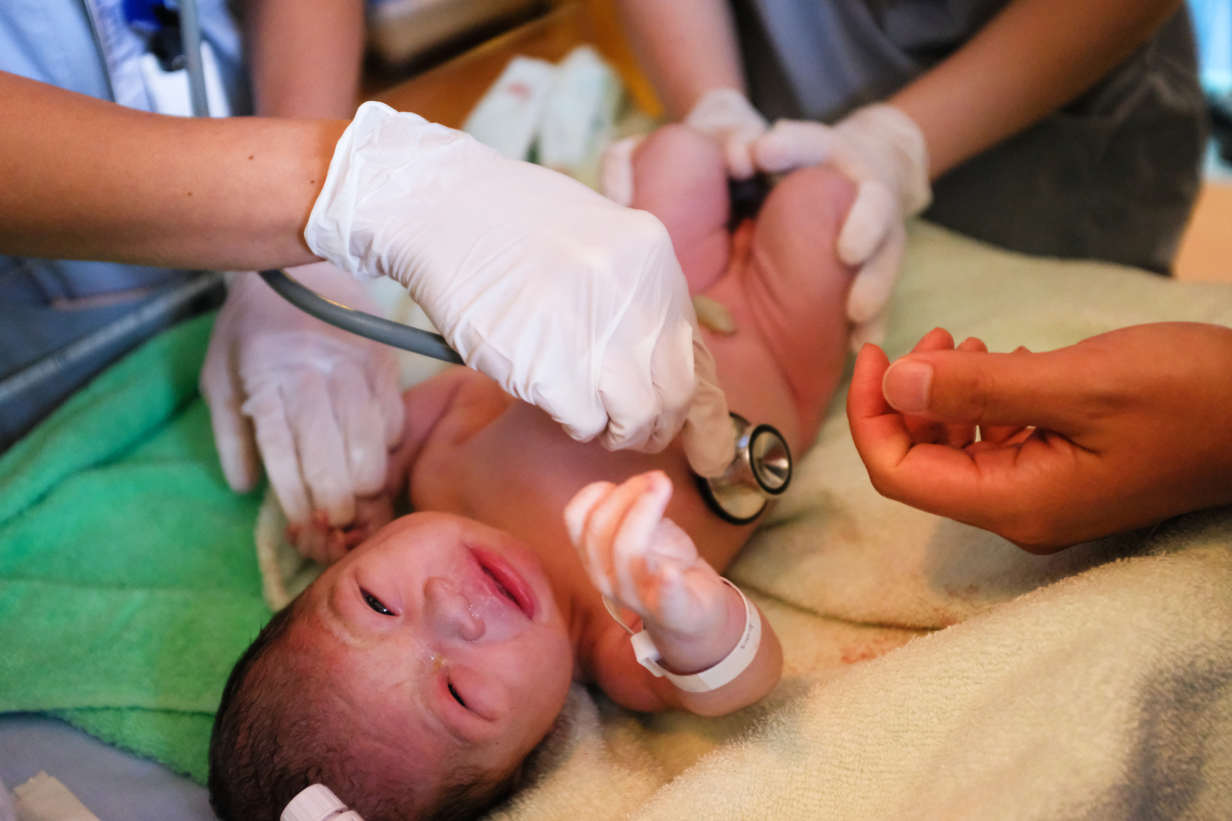 A baby's first exam on a warming table. Two sets of gloved hands--one holding baby's legs and one holding a stethoscope. 