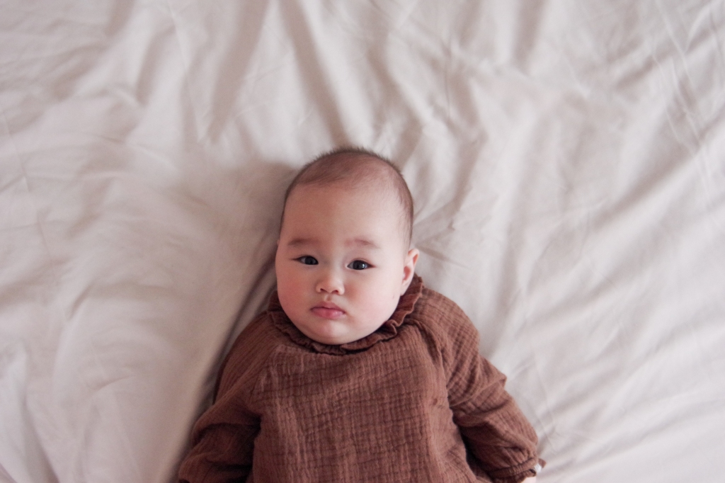 A 5 month baby in brown linen dress laying on a sheet looking up at the camera.