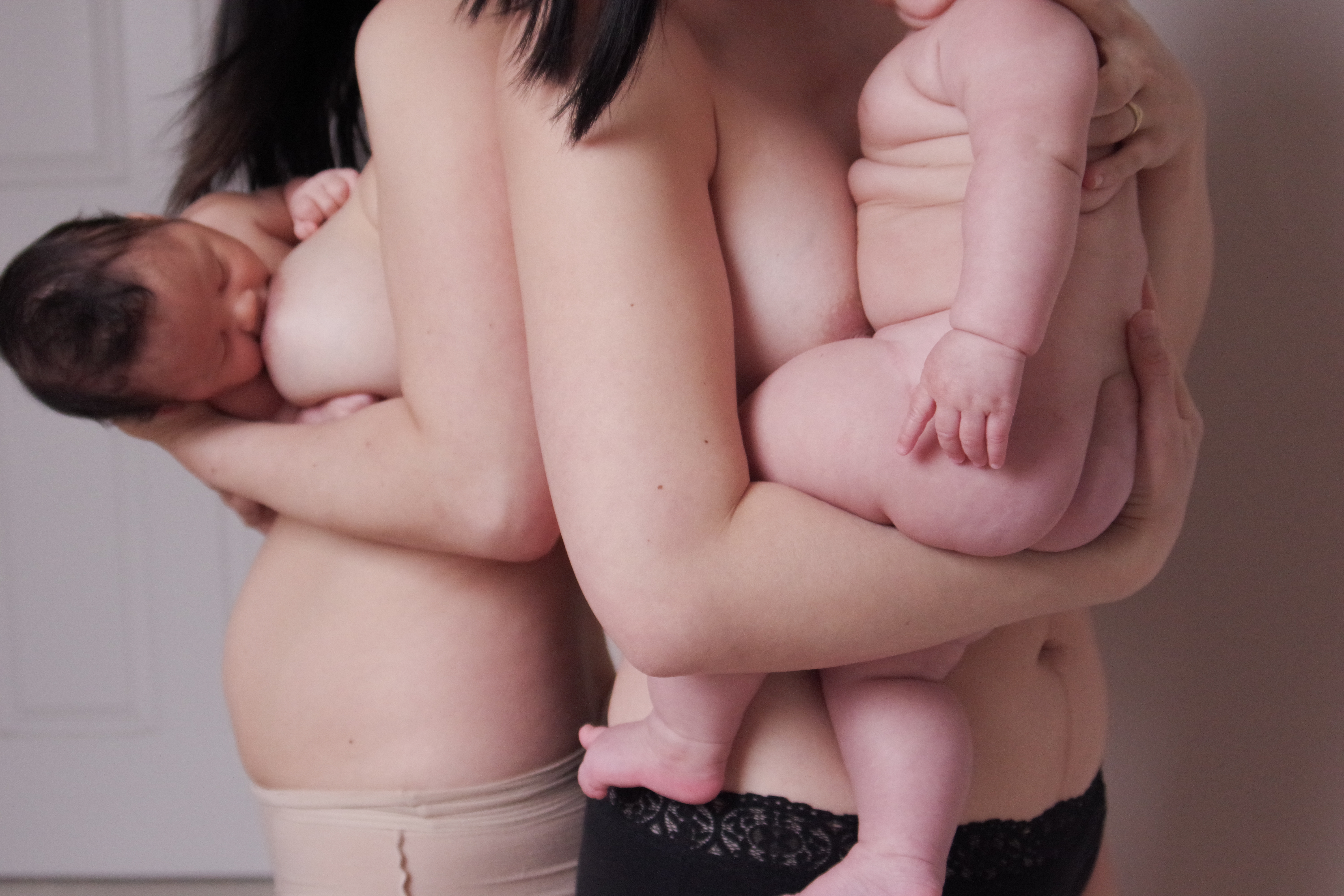 Two women stand naked holding their babies. One is chestfeeding while the other holds a 5 month old sitting up. The women's heads are cropped out, focussing on their bare skin torsos.
