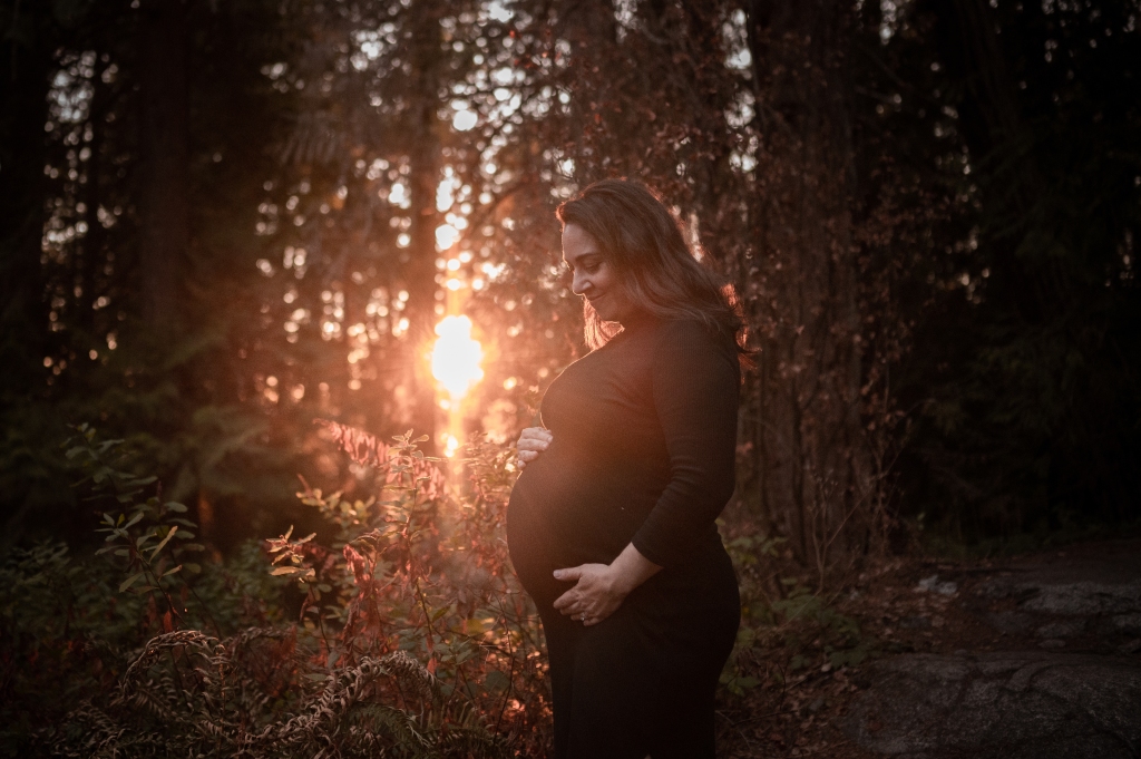 A backlit photograph of a woman holding her pregnant belly and looking down in a tight black dress. She is in a forest and the setting sun is shining through the trees in the background.